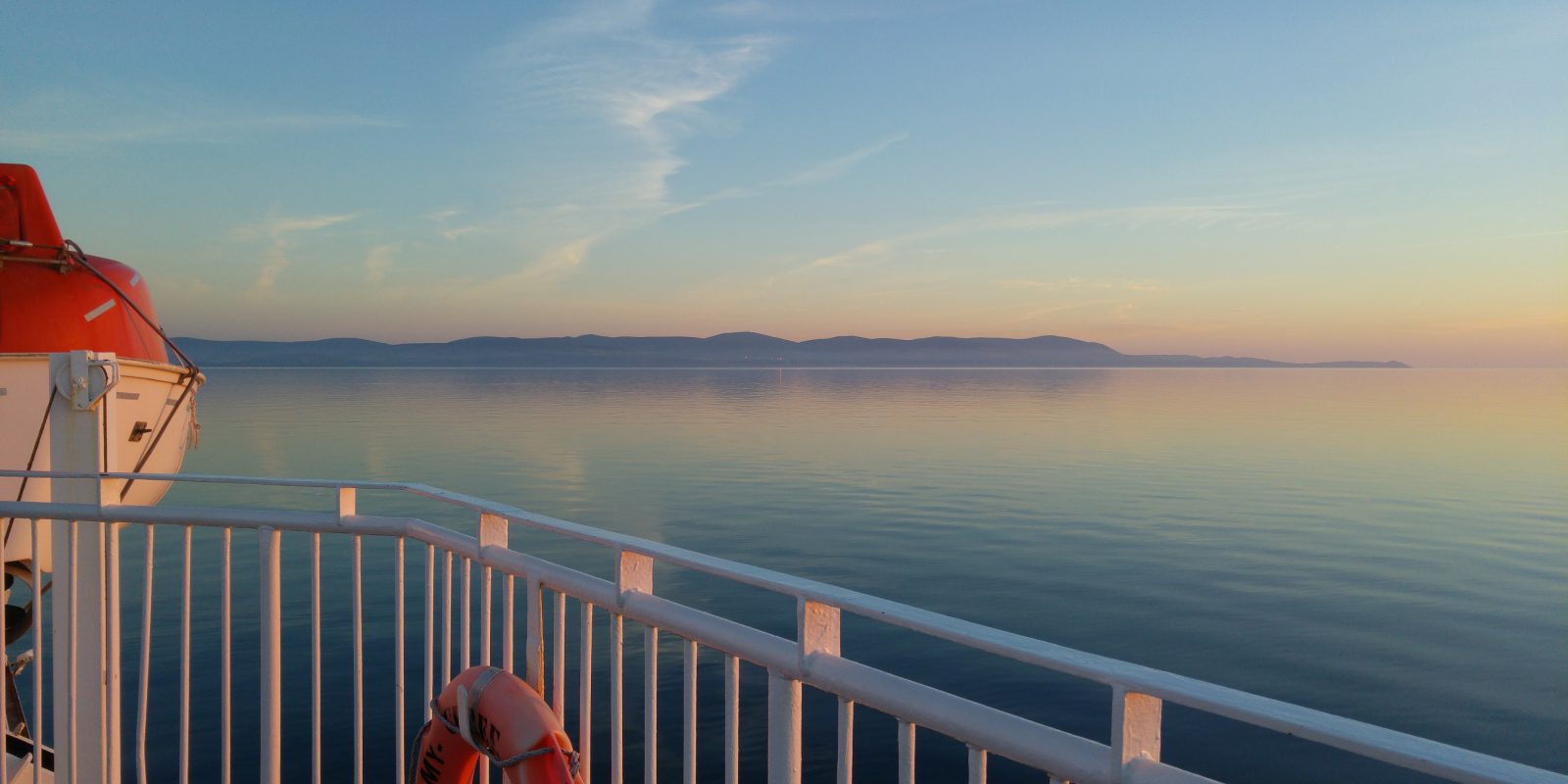 Looking out at dusk to sea with coastline in the background, and outside balcony of a ferry with lifebuoy and boat in foreground