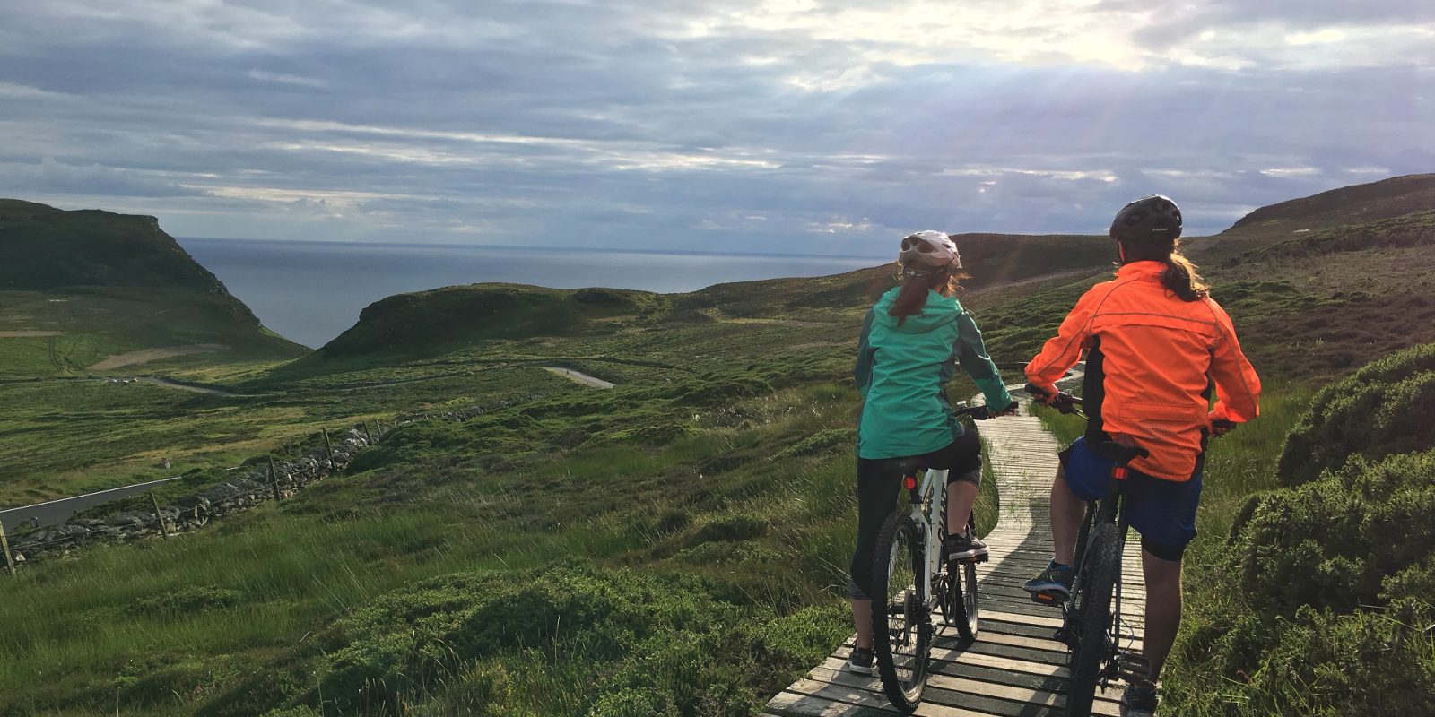 Two female cylist on their bikes standing on a wooden panelled path in a grassy mountain looking out to sea with the sun shining through the grey clouds