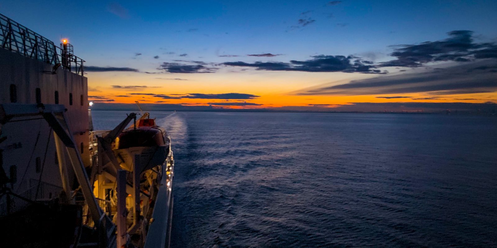 View from side of ferry, looking out from the back of the vessel to a sunset at sea with orange and blue sky