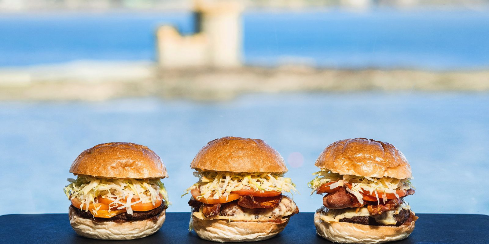 Marketing photo of 3 burgers side by side with the sea in the background