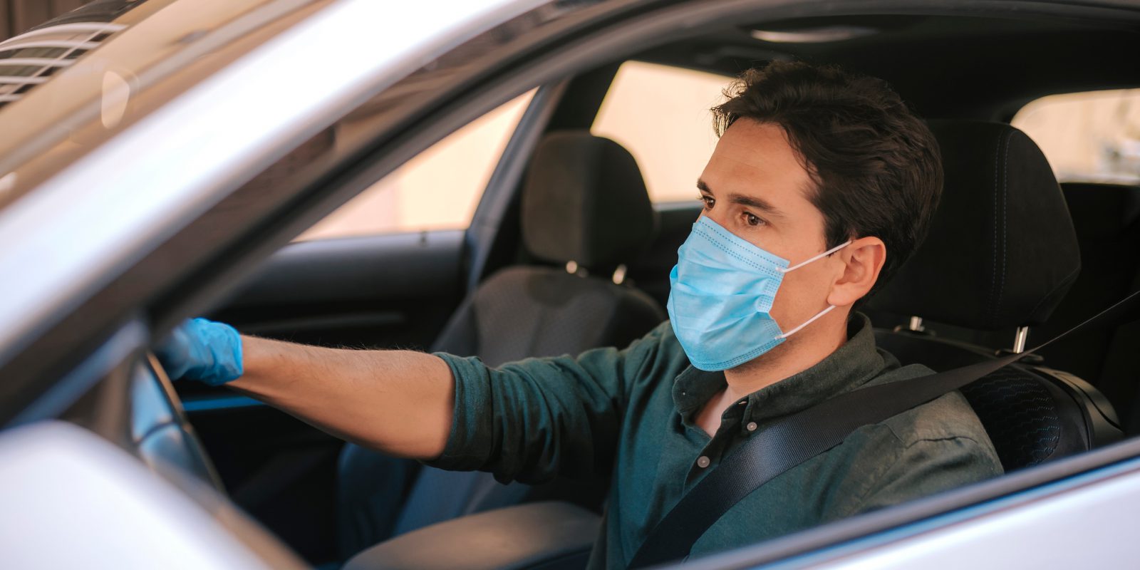 A man driving his silver car with the window down wearing a face mask