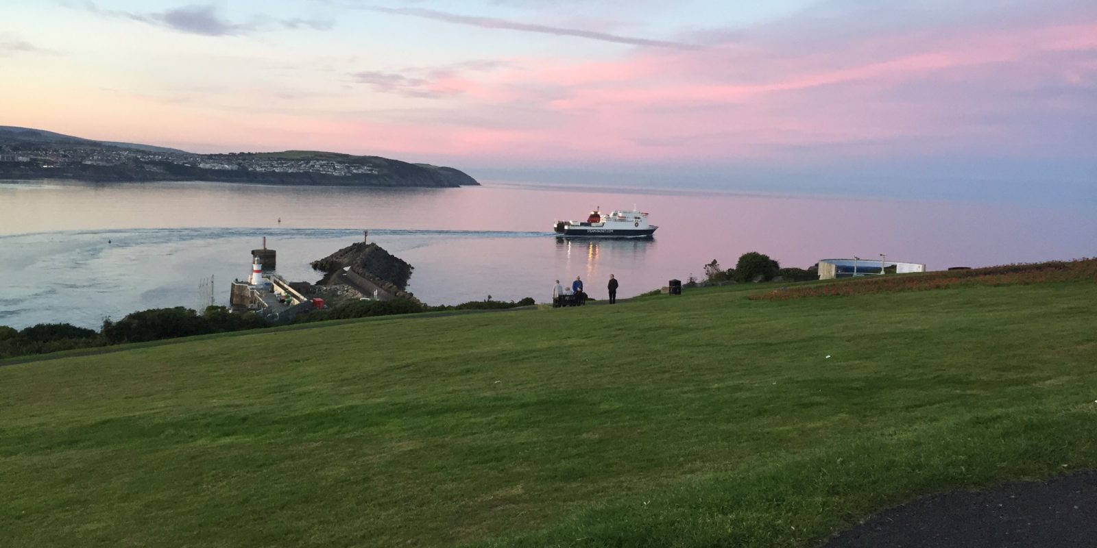 View from the grass on a hill looking out to a ferry going out to sea at sunset, pink skies with the coastline in the background