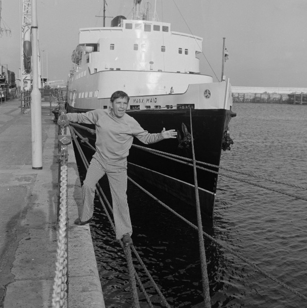 A black and white photo of Norman Wisdom posing happily next the Steam Packet Company vessel Manx Maid in Douglas Harbour in 1977